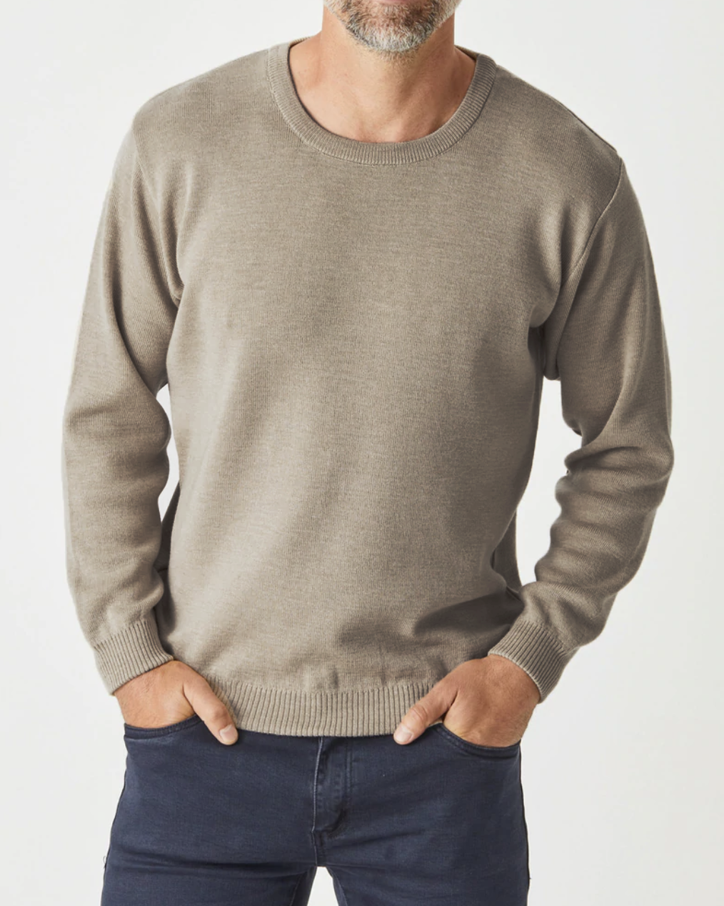 A person in an Aklanda Crew Pullover Australian Made, crafted from Merino Wool, paired with black pants. The beige jumper features long sleeves and a simple knitted texture as the individual stands against a white background with hands tucked into pant pockets.