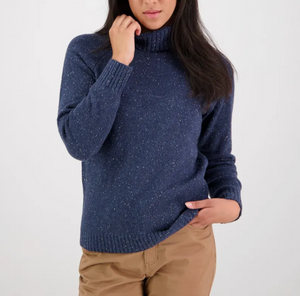 A person with long dark hair wears a navy Swanndri Lyttleton Roll Neck Jumper and light brown pants, standing against a plain white background. Their left hand is on their hip, while the right gently touches the sweater near the neck.