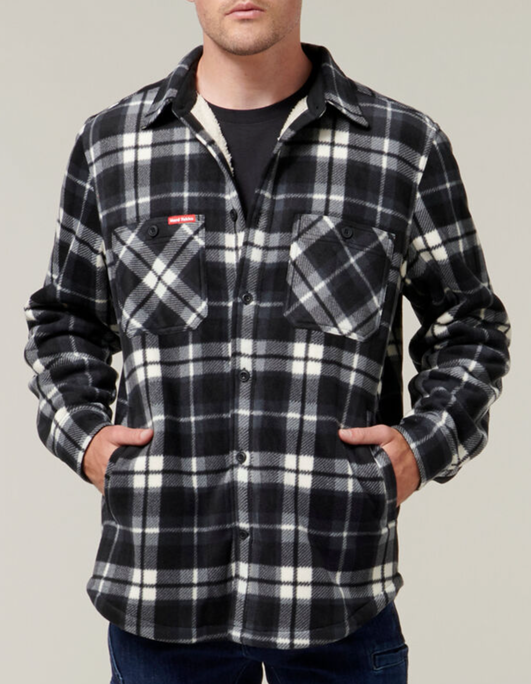 A person wearing the Hard Yakka Sherpa Bonded Fleece Jacket in black and white plaid, featuring sherpa lining and front pockets, stands with hands in the side pockets against a neutral background.