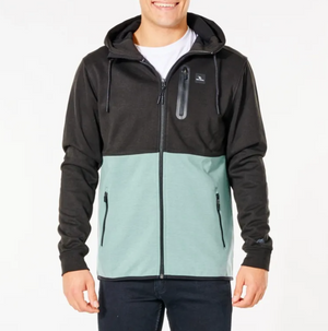 A person is wearing a Rip Curl Anti Series Departed Zip Thru hoodie, designed with a two-toned color scheme featuring a black upper section and a light green lower section. This Durable Water Resistant (DWR) hoodie includes a front zipper, two zippered pockets, and a small chest pocket with a zipper. The person is also dressed in dark pants and has a white shirt underneath.