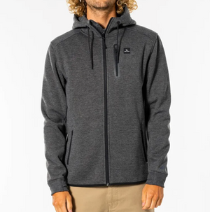 A person with curly hair is wearing a dark gray zip-up hoodie labeled as the Rip Curl Anti Series Departed Zip Thru, featuring Durable Water Resistant (DWR) technology. The hoodie has a hood and black drawstrings, paired with khaki pants. A small chest pocket with a zipper adorns the left side, and hands are relaxed at the sides. The background is plain white.