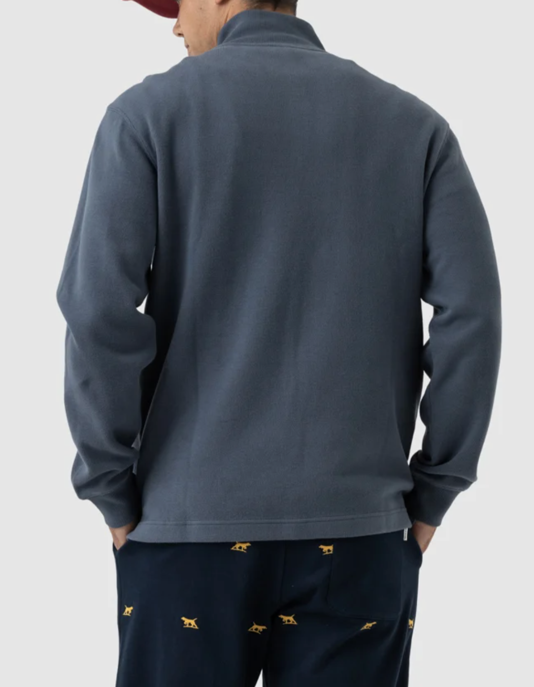 Rear view of a person wearing Rodd & Gunn Alton Ave Sweat, navy pants with a yellow animal pattern, and a red cap against a plain light gray background.
