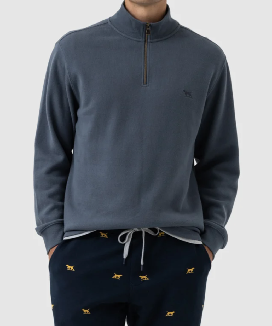 A person is wearing the Rodd & Gunn Alton Ave Sweat, a gray, 100% cotton pullover with a half-zip collar, paired with navy blue pants featuring small yellow embroidered dog patterns. The outfit exudes a casual and comfortable style against a plain, light-colored background.