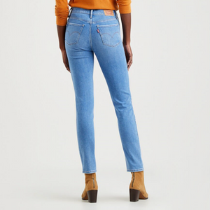 A person stands facing away, wearing a long-sleeve orange top, light blue Levis 312 Shaping Slim Jeans with a tummy-slimming panel, and tan heeled ankle boots against a plain white background. The jeans feature visible stitching and back pockets. The person's hands rest by their sides.
