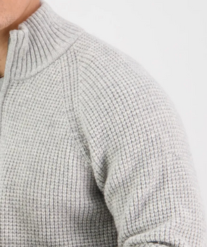 Close-up of a person wearing the Swanndri Chalky Island Waffle Jumper, a light gray knit sweater with a textured waffle pattern and high collar. The fitted design showcases a partially visible zipper at the collar against a plain white background.