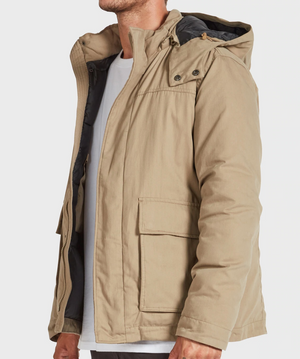 A man wears the Academy Brand Miller Jacket—a light brown hooded winter layer over a white shirt. Shown from the side, his face is partially out of view. The jacket features large front pockets and a zippered button flap.