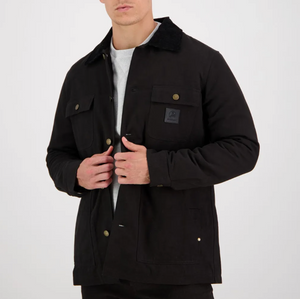 A person in a Swanndri Barrytown Canvas Work Jacket, featuring buttoned pockets and a black collar, is standing against a plain white background. They are wearing a light gray shirt underneath and are adjusting the jacket with their hands. The jacket, which features quilted lining, has a patch on the left chest pocket.