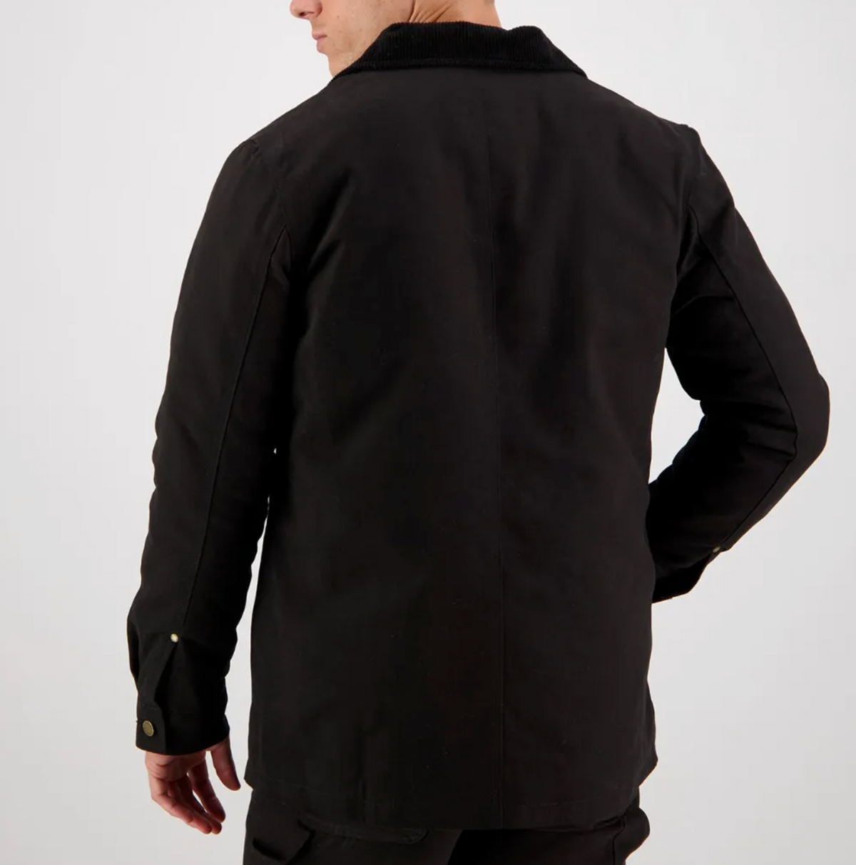 A person wearing a Swanndri Barrytown Canvas Work Jacket stands with their back to the camera, their right hand resting on their hip. The jacket features buttoned cuffs and a textured collar, with a quilted lining subtly peeking out. The plain, light-colored background accentuates the dark clothing.