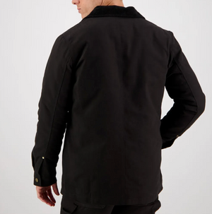 A person wearing a Swanndri Barrytown Canvas Work Jacket stands with their back to the camera, their right hand resting on their hip. The jacket features buttoned cuffs and a textured collar, with a quilted lining subtly peeking out. The plain, light-colored background accentuates the dark clothing.