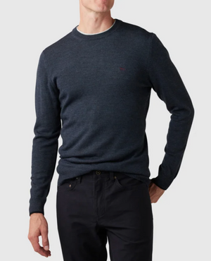 A person wearing a Rodd & Gunn Abberfield Knit sweater in dark blue stands against a plain background, paired with dark pants. The Merino wool sweater features long sleeves and a small chest logo. They have one hand in their pocket and are turned slightly to the side.