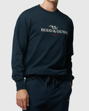 A person wearing a Rodd & Gunn Grenada North Sweat in dark blue, featuring the "Rodd & Gunn Sport" logo on the front, stands against a plain background. The 100% cotton sweatshirt has long sleeves and a round neckline, paired with matching dark blue pants that have a drawstring waist, perfect for a comfortable sweat.