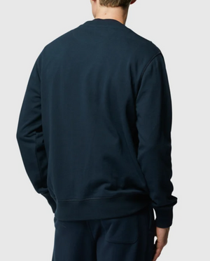 A person wearing a dark blue Rodd & Gunn Grenada North Sweat and matching pants stands with their back to the camera. The background is plain white, and their head is slightly turned to the left, showcasing the comfortable sweat ensemble made from 100% cotton.