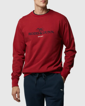 A man models the Rodd & Gunn Grenada North Sweat, a 100% cotton red crew neck with "RODD & GUNN SPORT" embroidered on the front, paired with navy joggers. The image shows him from the waist up against a plain background.