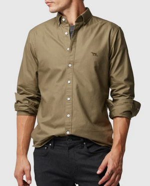 A person models a Rodd & Gunn Oxford Pd Sports Fit shirt in khaki, sleeves partially rolled up, with an embroidered chest logo. Made from 100% cotton, it pairs well with fitted black jeans against a plain, neutral background.