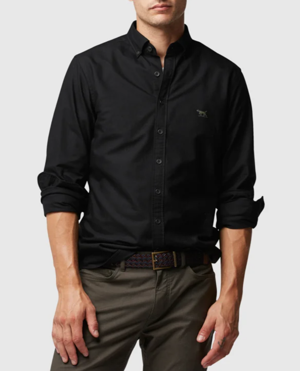 A person is sporting a stylish Rodd & Gunn Oxford Pd Sports Fit Shirt made of 100% cotton, with sleeves partially rolled up, paired with olive-green pants. The long-sleeved black button-up shirt features a small embroidered logo on the chest, though the person's head is not visible in the image.