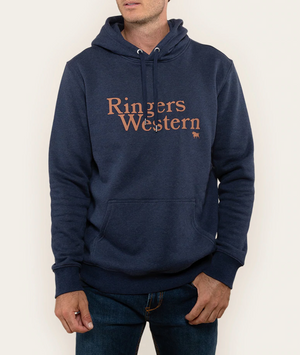 A person is wearing a Ringers Western Lodge Hoodie in dark blue, featuring the words "Ringers Western" printed in orange on the front. The relaxed-fit, comfortable hoodie pairs perfectly with their dark blue jeans. They have their right hand tucked into the hoodie’s front pocket while standing against an off-white background.