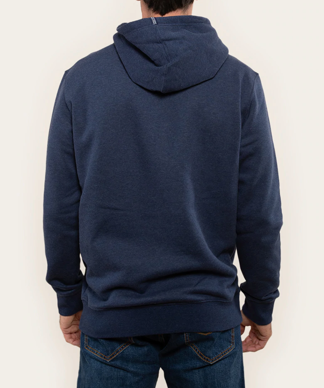 Revised Sentence: Rear view of a person wearing a dark blue Ringers Western Lodge Hoodie and blue jeans. The individual's arms are relaxed by their sides, and the hood of the comfortable sweatshirt is down. The background is a plain, light beige color.