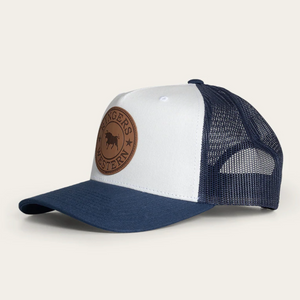 The Ringers Western Whitby Trucker Cap features a white front panel with a brown leather patch displaying a buffalo and stars, a curved brim, and blue mesh back panels for classic appeal.