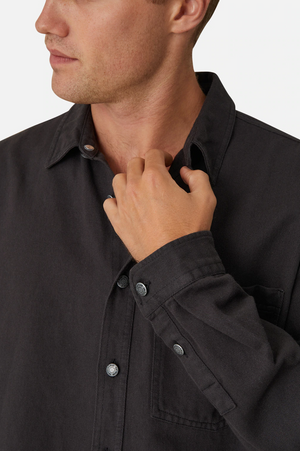 A person with short hair, partially visible, adjusts the collar of their stylish Industrie The Portland Shirt. The charcoal-colored cotton/linen blend shirt features a front pocket, and the person's face is turned slightly to the side, looking out of view.