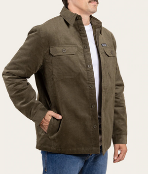 A person in a Ringers Western Ashwell olive-green overshirt with chest pockets, layered over a white shirt and blue jeans, stands with one hand in their pocket. The neutral background enhances the look.