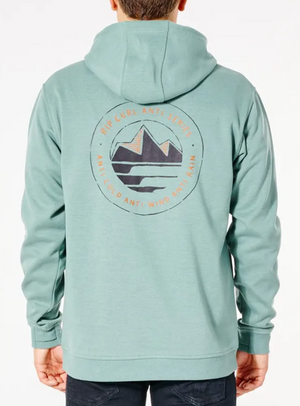 A person wears a Rip Curl Anti Series Journey Hood, a teal hoodie with a mountain graphic on the back, layered for winter. This durable, water-resistant design highlights protection against cold, wind, and rain.