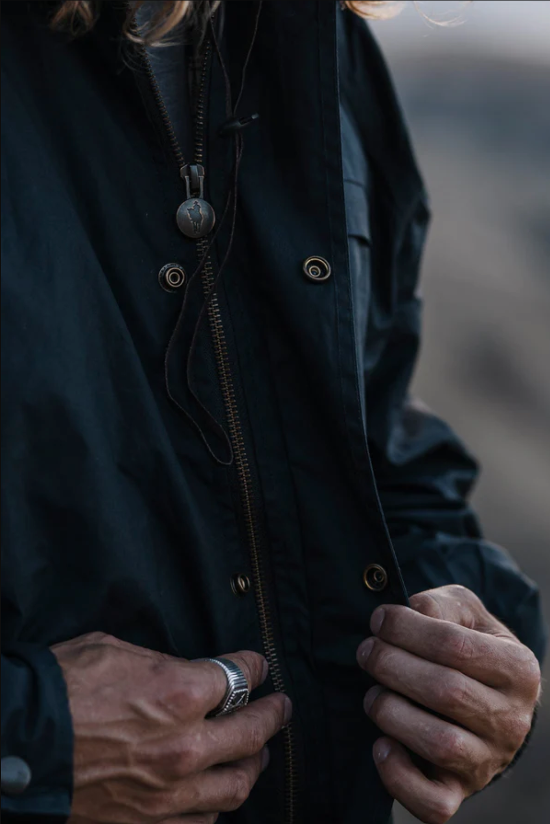 A person zips up their dark Outback Burke Jacket. Long-haired and wearing a ring, they sport a pendant on a cord, adding an air of mystery to their look. The blurred background enhances the allure of their enigmatic presence.