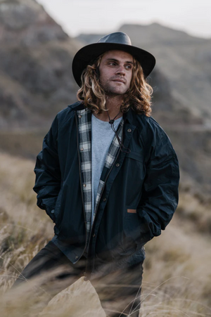 A person with long hair stands outdoors on grassy hills, wearing a wide-brimmed hat, the Outback Burke Jacket, and a plaid shirt. They have their hands in their pockets and look to the side.