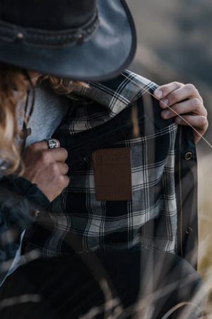 A person with long hair, wearing a dark hat and ring, holds open an Outback Burke Jacket showing its plaid flannel lining and brown label. The background features blurred natural tones that enhance the charm of this Australian waxed cotton waterproof jacket.