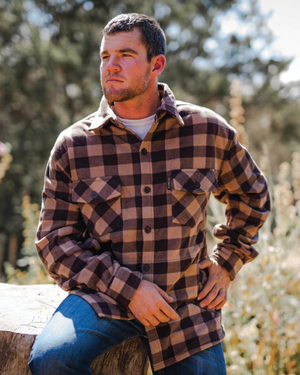 A man outdoors stands with one hand on his hip in an Outback Mens Big Shirt, a mid-weight garment featuring a cozy fleece design perfect for cool weather. Leaning against a large log, he looks to the right as sunlight filters through the leafy backdrop.