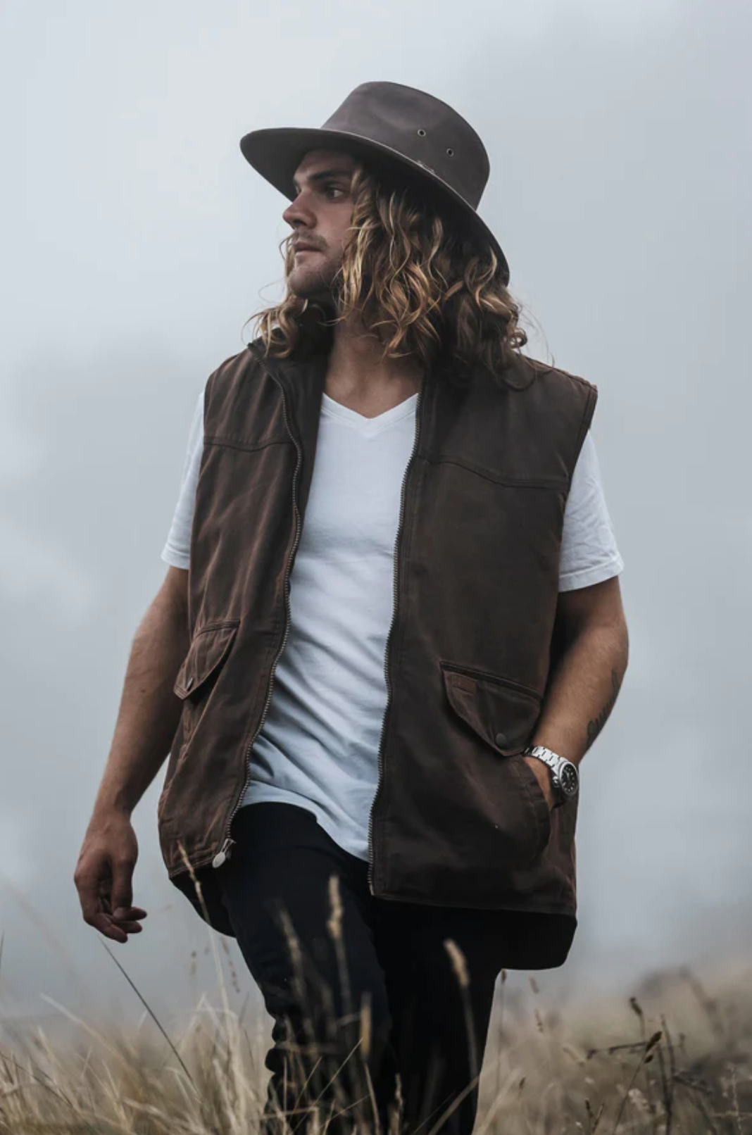 A man with long hair and a beard stands in a foggy field. He wears a wide-brimmed hat, an Outback Landsman Vest over a white t-shirt, and black pants. The water-resistant vest is machine washable. The man looks off into the distance with his hands in the pockets of his vest.