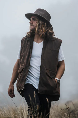 A man with long hair and a beard stands in a foggy field. He wears a wide-brimmed hat, an Outback Landsman Vest over a white t-shirt, and black pants. The water-resistant vest is machine washable. The man looks off into the distance with his hands in the pockets of his vest.