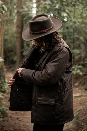 A person with long hair, wearing an Outback Ranchers Jacket, features a dark brown, hard-wearing exterior and a polar fleece lining. They also have on a wide-brimmed hat with a reddish band. They stand in a forest, adjusting or inspecting the jacket's inside pocket amid trees and green foliage.