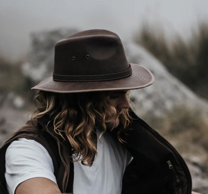 A person with long hair, dressed in a white shirt and brown vest, sits outdoors wearing the Outback Raven Hat. The wide-brimmed hat offers UPF protection and moisture-wicking properties, ensuring comfort as they look downward against a foggy backdrop of blurred rocks and grass.