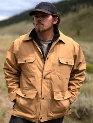 A man with a beard stands outdoors wearing an Outback Thomas Canvas Jacket, which is made of beige Heavy Duty Cotton Canvas, and a black cap. His hands are in his jacket pockets. The background features a blurred natural landscape with hills and green vegetation.