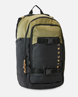The Rip Curl Posse 33L Overland Backpack is a stylish two-tone accessory with an olive green upper section and a black lower section. Crafted from recycled polyester, it includes several zippered compartments, an internal laptop section, adjustable straps with buckles, and the "RIP CURL" brand printed vertically in orange on the front.