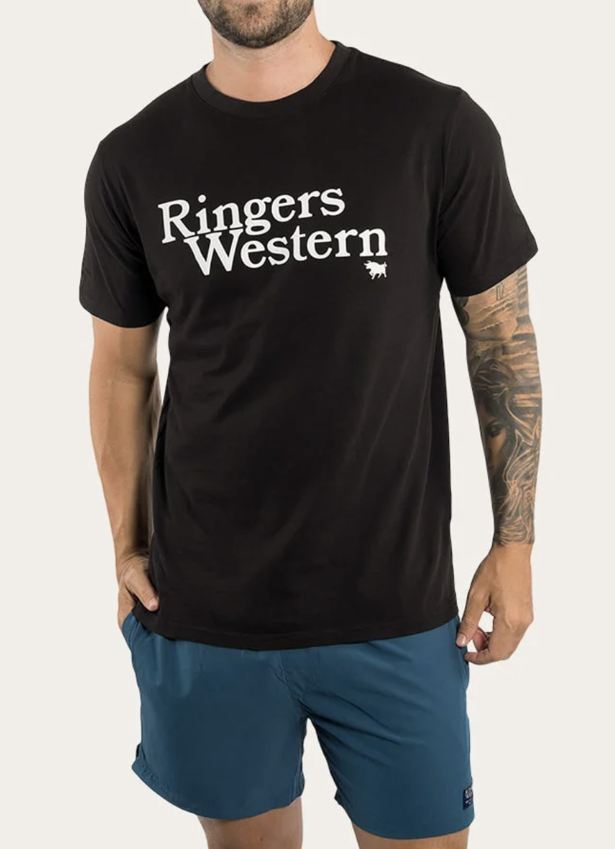 A man wearing the Ringers Western The Lodge Classic Fit T Shirt, made from 100% cotton and featuring white text with a small steer head logo, stands in blue shorts with his tattooed left arm visible and one hand in his pocket.