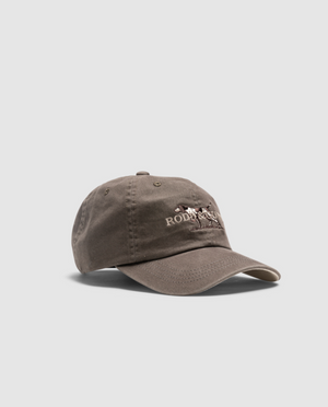 The Rodd & Gunn Double Barrel Cap is brown with a slightly worn texture, crafted from Heavy Duty Cotton Twill. It features an embroidered design of a horse and rider, accompanied by the text “Polo” and “Ralph Lauren” in white. An adjustable strap ensures the perfect fit, all set against a plain, light grey background.