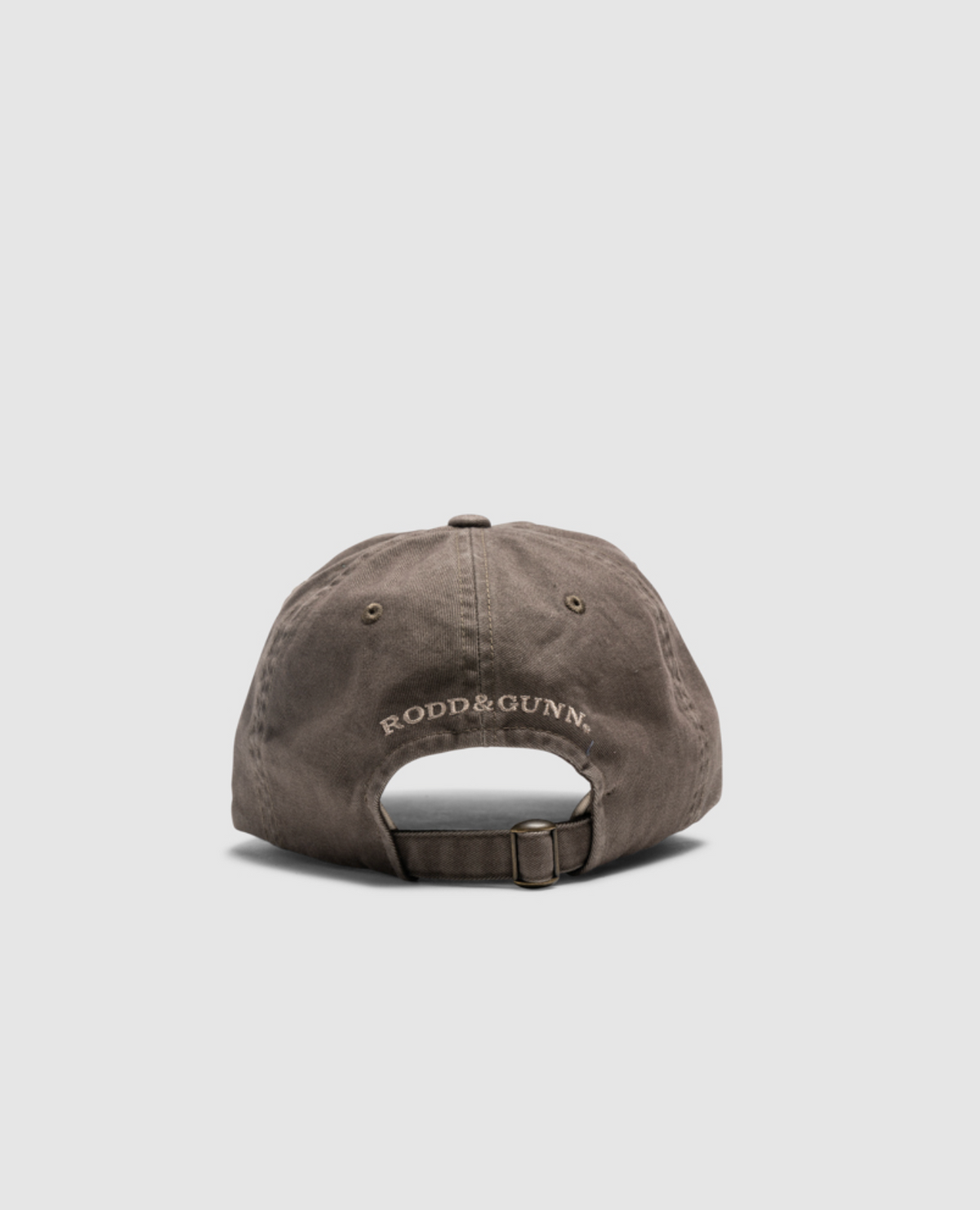 A brown Rodd & Gunn Double Barrel Cap, made from heavy-duty cotton twill, is displayed from the back. The brand's name is embroidered with white thread above the adjustable strap on the rear panel. The background is a plain light grey.
