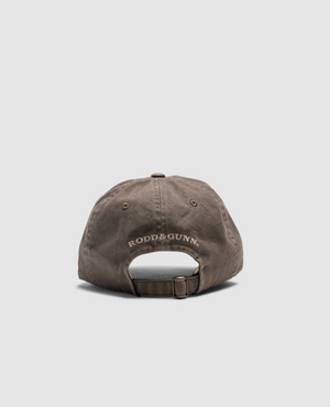 A brown Rodd & Gunn Double Barrel Cap, made from heavy-duty cotton twill, is displayed from the back. The brand's name is embroidered with white thread above the adjustable strap on the rear panel. The background is a plain light grey.
