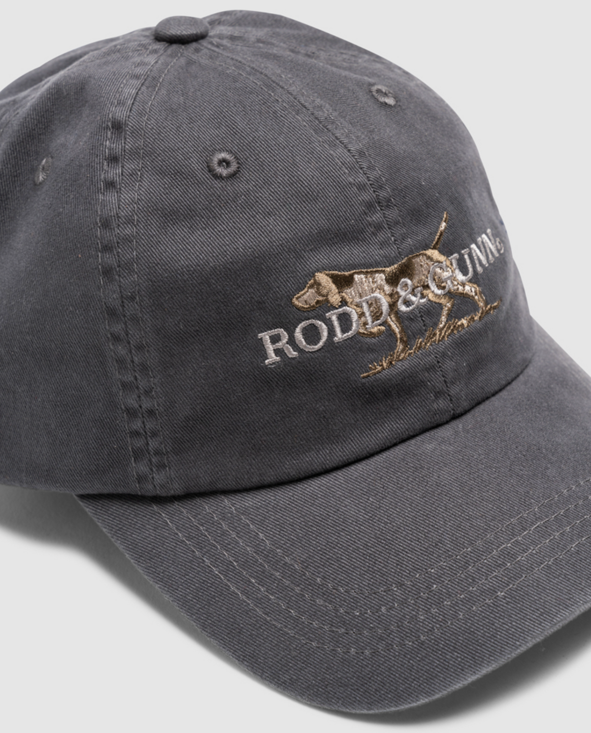Dark gray Rodd & Gunn Double Barrel Cap in cotton twill, features an embroidered dog and "RODD & GUNN" text on the front, tilted slightly right to reveal its curved brim and adjustable back strap.
