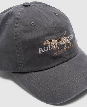 Dark gray Rodd & Gunn Double Barrel Cap in cotton twill, features an embroidered dog and "RODD & GUNN" text on the front, tilted slightly right to reveal its curved brim and adjustable back strap.