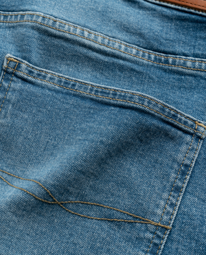 Close-up of a Rodd & Gunn Stephens Bay Summer Short pocket in blue denim with visible white stitching, showcasing the stretch fit. The fabric texture is highlighted, and part of a brown leather belt loops neatly at the top edge of this mid-rise waist image.