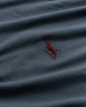 A close-up image of the Rodd & Gunn The Gunn T-Shirt, crafted from Australian cotton in a deep blue hue, features an embroidered logo of a red dog standing on its hind legs, seemingly holding or leaning on a small object. The fabric appears slightly creased.