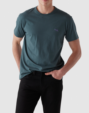 Someone wears a teal Rodd & Gunn The Gunn T-Shirt made from premium Australian Cotton, featuring an embroidered animal logo on the chest. Paired with black pants on a white background, its sports fit lets one hand rest in a pocket while the other is by their side. Their face isn't shown.