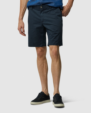 A person dons dark navy Rodd & Gunn The Peaks Custom Shorts with a stretch fit and black sneakers with white soles. They're against a plain white backdrop, their left hand in their pocket, and only their lower half is visible.