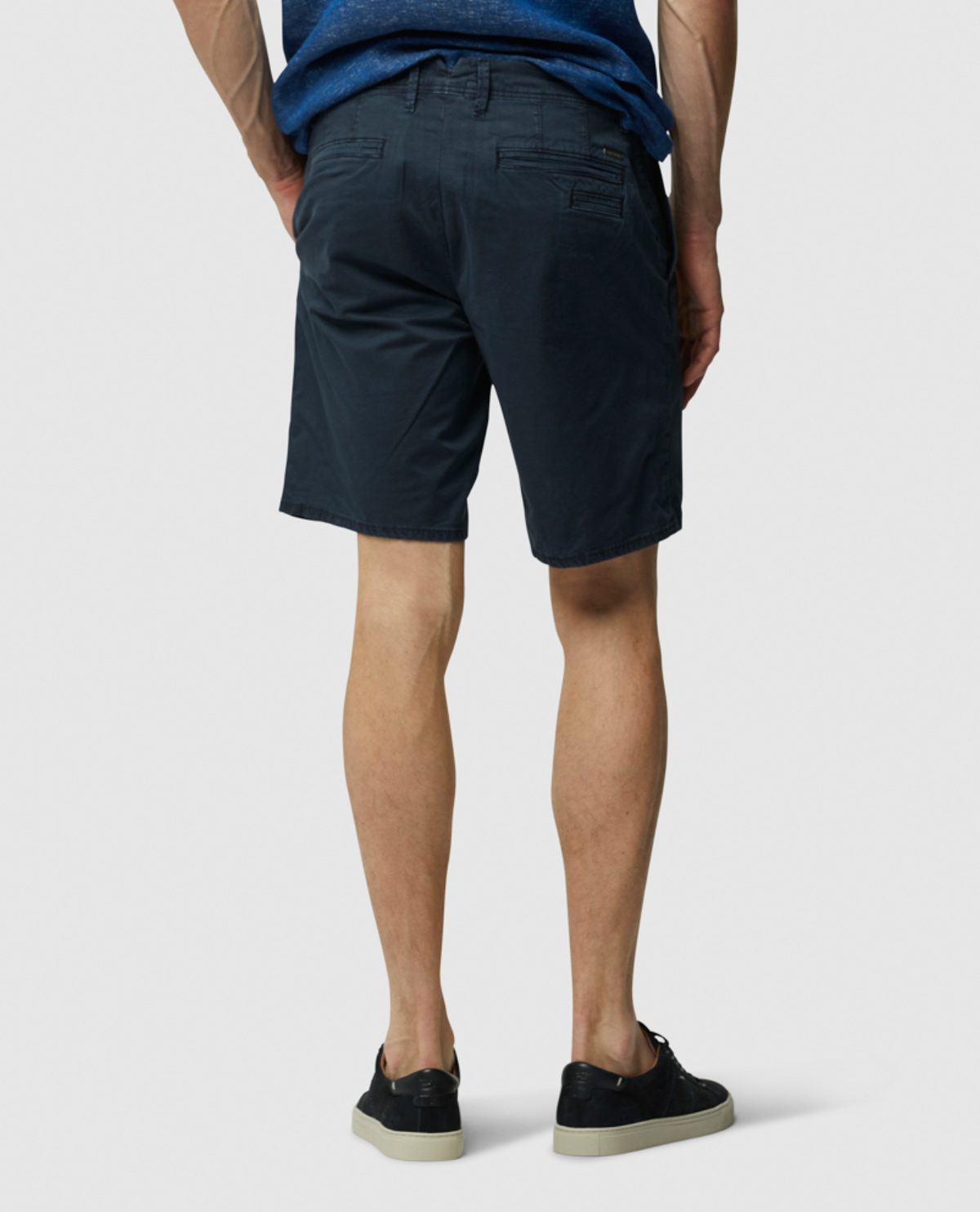 A person, viewed from behind against a gray background, is wearing a blue stretch-fit shirt, Rodd & Gunn The Peaks Custom Short in dark navy, and black sneakers with beige soles.