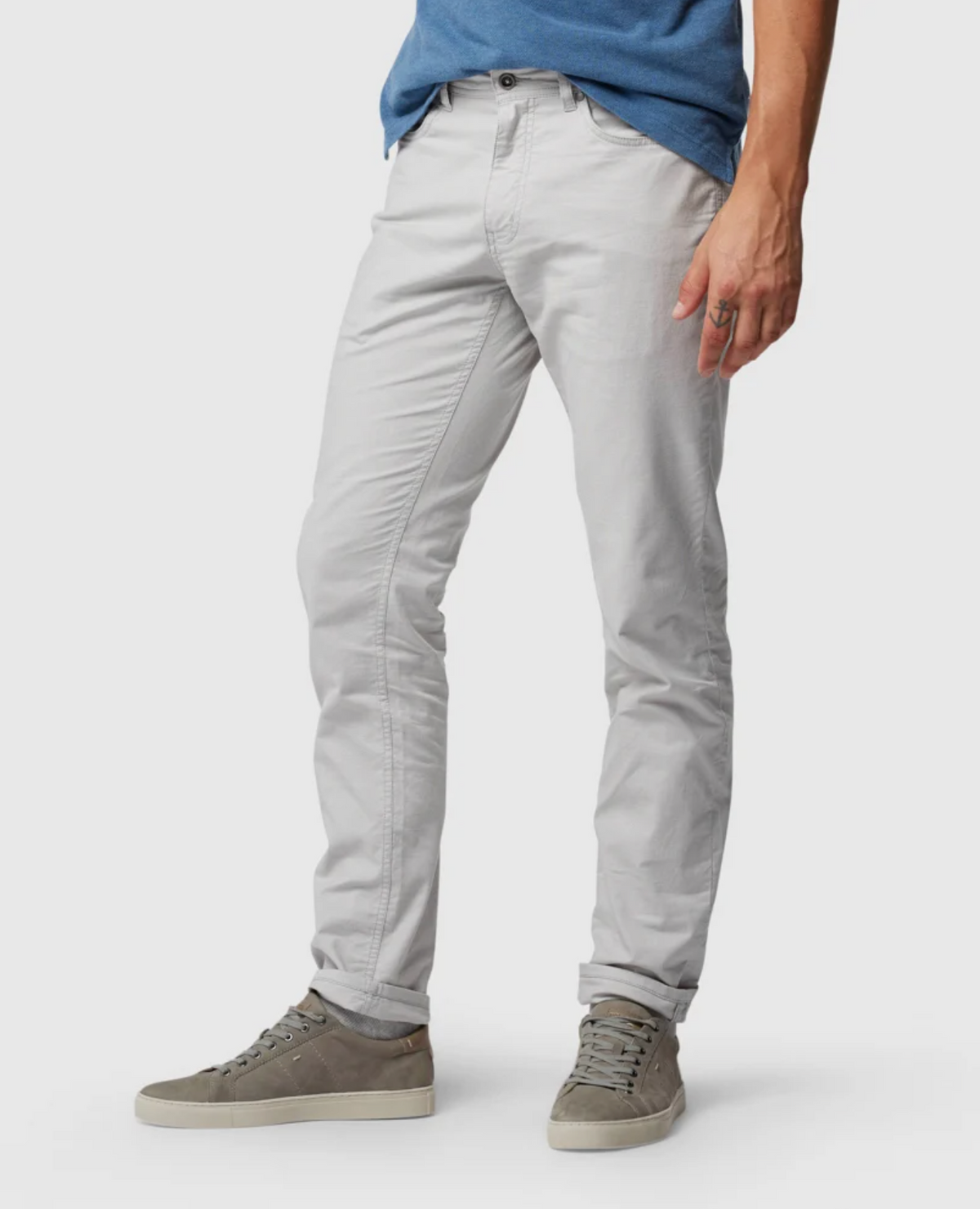 A person wearing Rodd & Gunn Straight Fit Jeans with a YKK zip fly, a blue shirt partially visible, and grey suede sneakers is posed in a relaxed manner with one hand resting by their side. The photo captures the ensemble from the waist down against a plain white background.