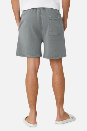 A person in a white shirt, Industrie The Del Sur Track Short (gray, oversized fit, elastic waist, back pocket), and white slippers stands facing away with hands in side pockets. The background is plain white.
