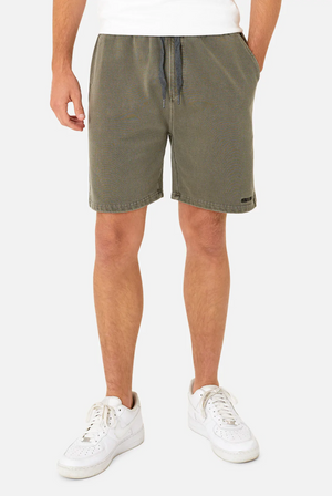 A person confidently sports the olive green Industrie The Del Sur Track Short, featuring an elastic waist and drawstring, paired with white sneakers. With hands in pockets and a small tag on the left leg, they stand against a plain white background.