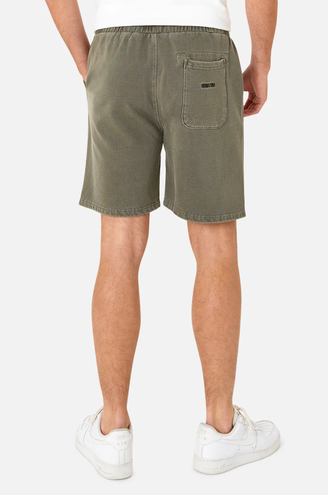 A person is seen from the back, wearing Industrie The Del Sur Track Short in olive green with an oversized fit and white sneakers. The shorts have an elastic waist and a right-side back pocket with text. They stand against a plain white background.